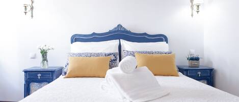 A neatly made bed with white and blue bedding, two pillows, and a rolled towel. There are two bedside tables with lamps and a vase of flowers on one.