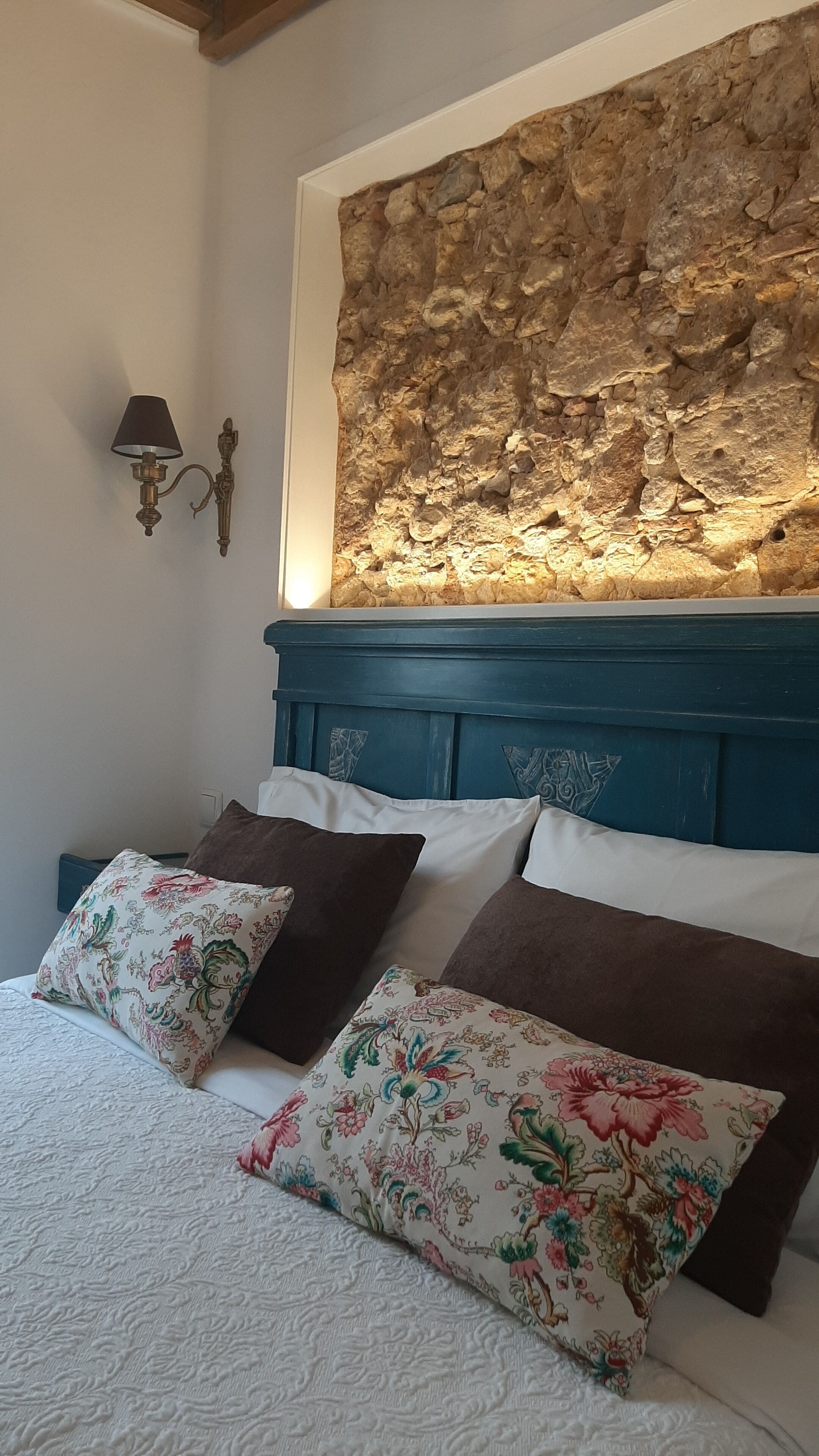 Un dormitorio con pared de piedra, cabecero azul y almohadas estampadas con motivos florales.