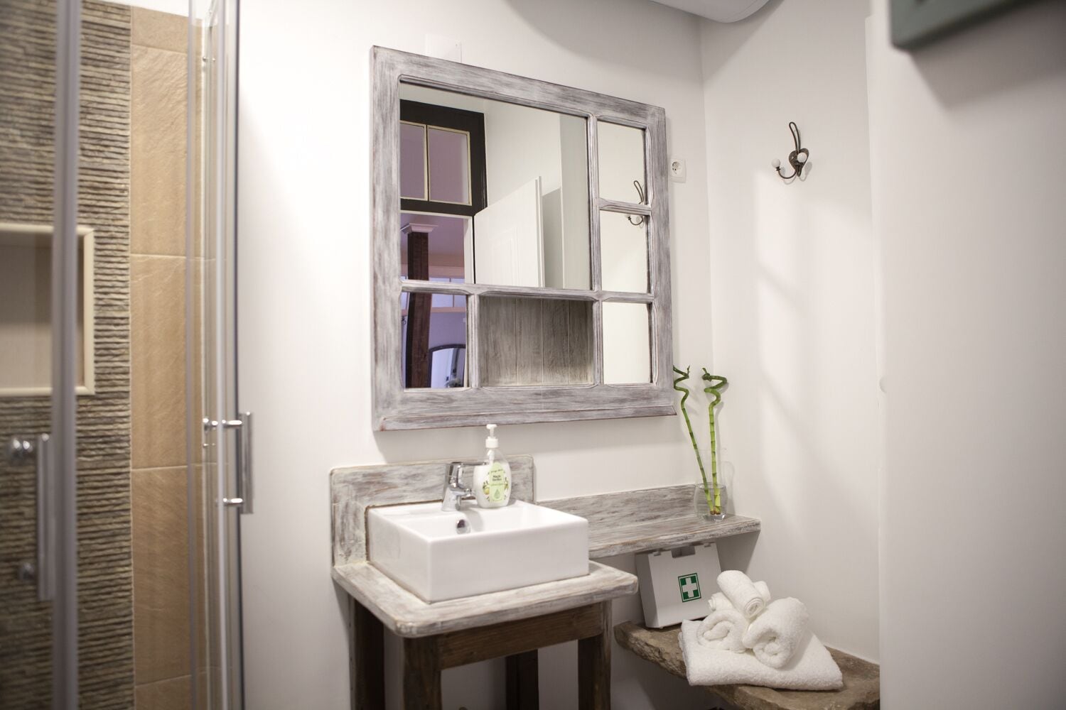 A bathroom with a white sink, a wooden vanity, a mirror with a distressed frame, a glass vase with green plants, and a first aid kit on the vanity.