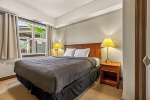 A neatly made bed with a headboard, two bedside lamps, a nightstand, and a window with curtains. - 1 Bedroom Plus Studio Lock-Off Unit. AC (Fernie)