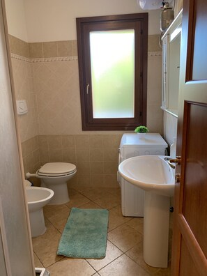 A small bathroom with a toilet, bidet, and sink, a window with frosted glass, and a green towel on the floor.