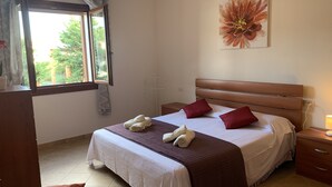 Une chambre à coucher bien rangée, avec un lit, des oreillers, une table de chevet, une commode, une fenêtre avec des rideaux et un tableau représentant des fleurs accroché au mur.