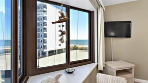 Une pièce dotée d’une grande fenêtre donnant sur une plage et un immeuble, d’un téléviseur à écran plat fixé au mur, d’un fauteuil en osier et d’une petite table agrémentée d’objets décoratifs.