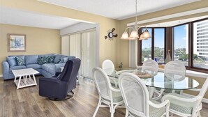 Un salon moderne avec une table à manger en verre, des chaises en osier blanches, un canapé bleu et une grande fenêtre donnant sur la ville.