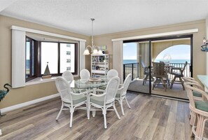 A dining area with a glass table and white wicker chairs, a bookshelf, and a view of the ocean through large sliding glass doors. - Huge ocean front balcony in this 2 Bedroom condo in the 9400 Building! (Ocean City)