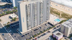 A high-rise hotel with multiple floors, a parking lot, and a beach view.