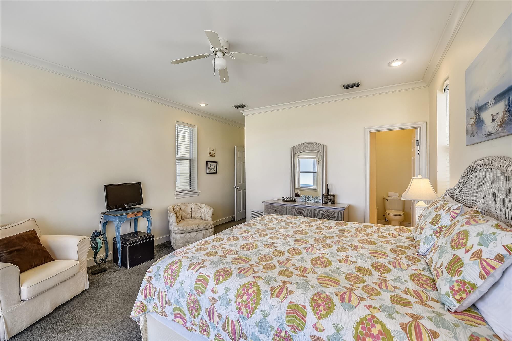 A bedroom with a large bed, a ceiling fan, a dresser, a chair, a television, and a window with blinds.