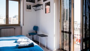 Een slaapkamer met een bed, een stoel, een tafeltje en een raam met uitzicht op gebouwen.