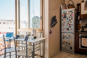 Une cuisine avec un réfrigérateur recouvert d’aimants, une fenêtre avec des stores et une table avec des chaises.