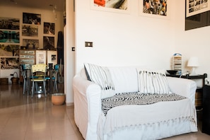Un salon avec un canapé blanc, un plaid à motifs, un coin repas comprenant des chaises et une table, et un mur décoré de photographies.