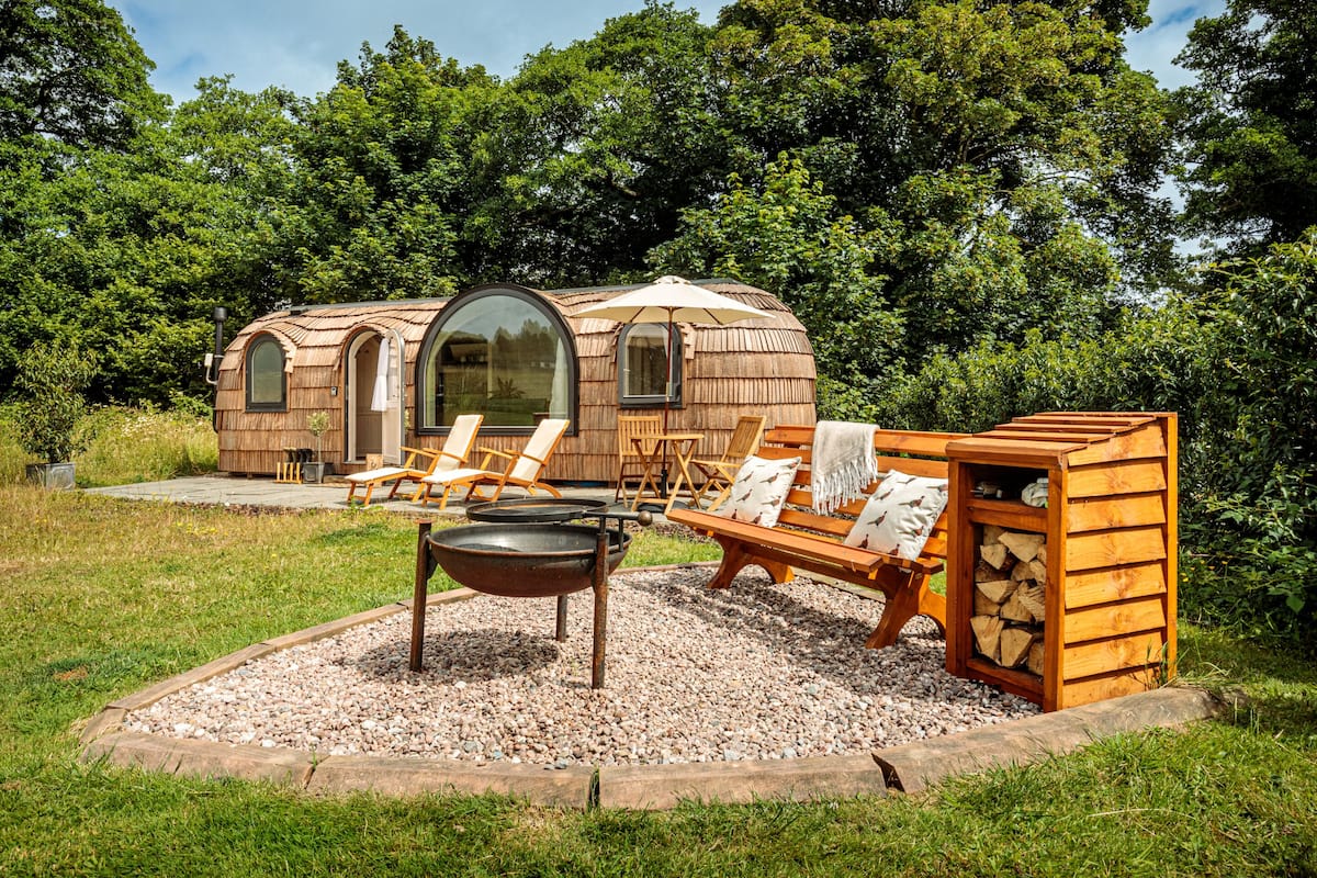 A wooden cabin with a circular window, outdoor seating, and a fire pit.