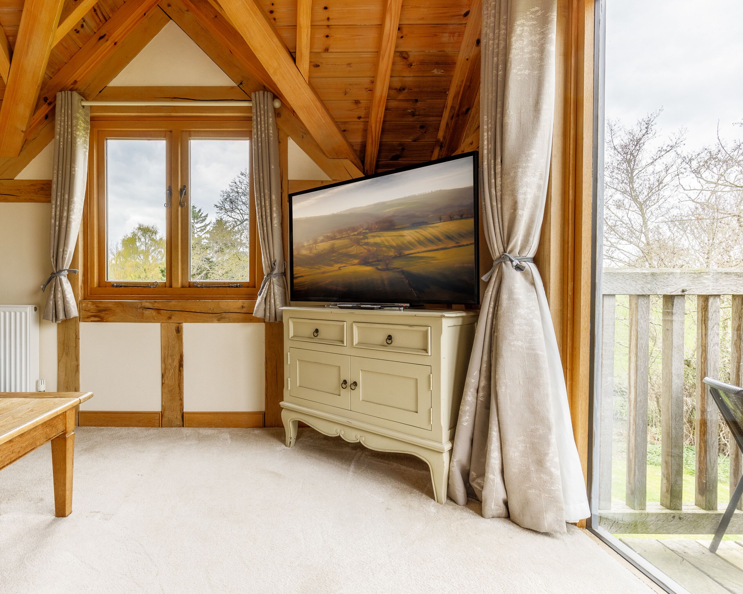 Una habitación acogedora con techo de madera, un ventanal con cortinas, un televisor de pantalla plana sobre un mueble color crema y vistas a árboles.