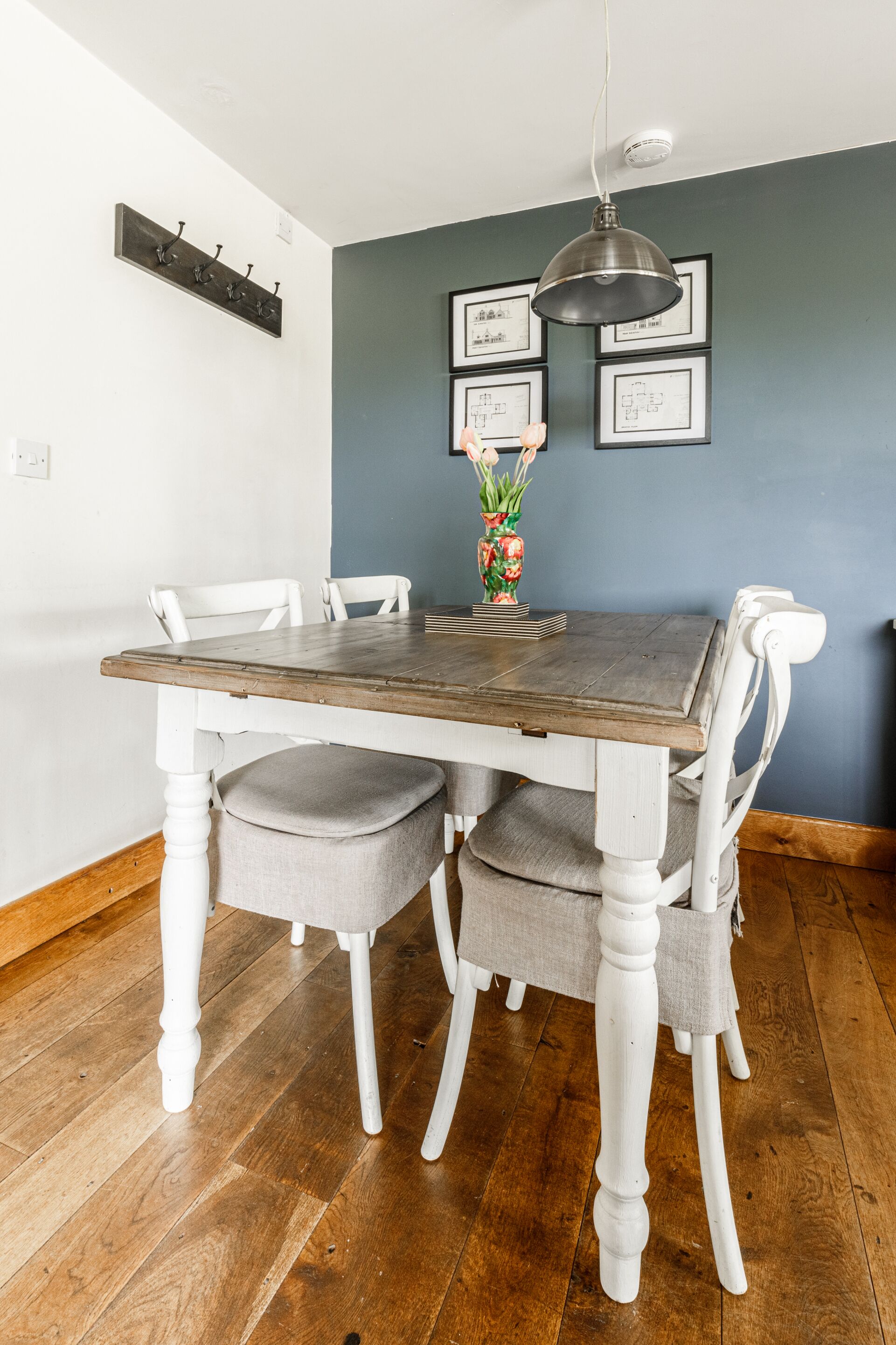 Una mesa de comedor con sillas, un jarrón con flores y dibujos arquitectónicos enmarcados en la pared.