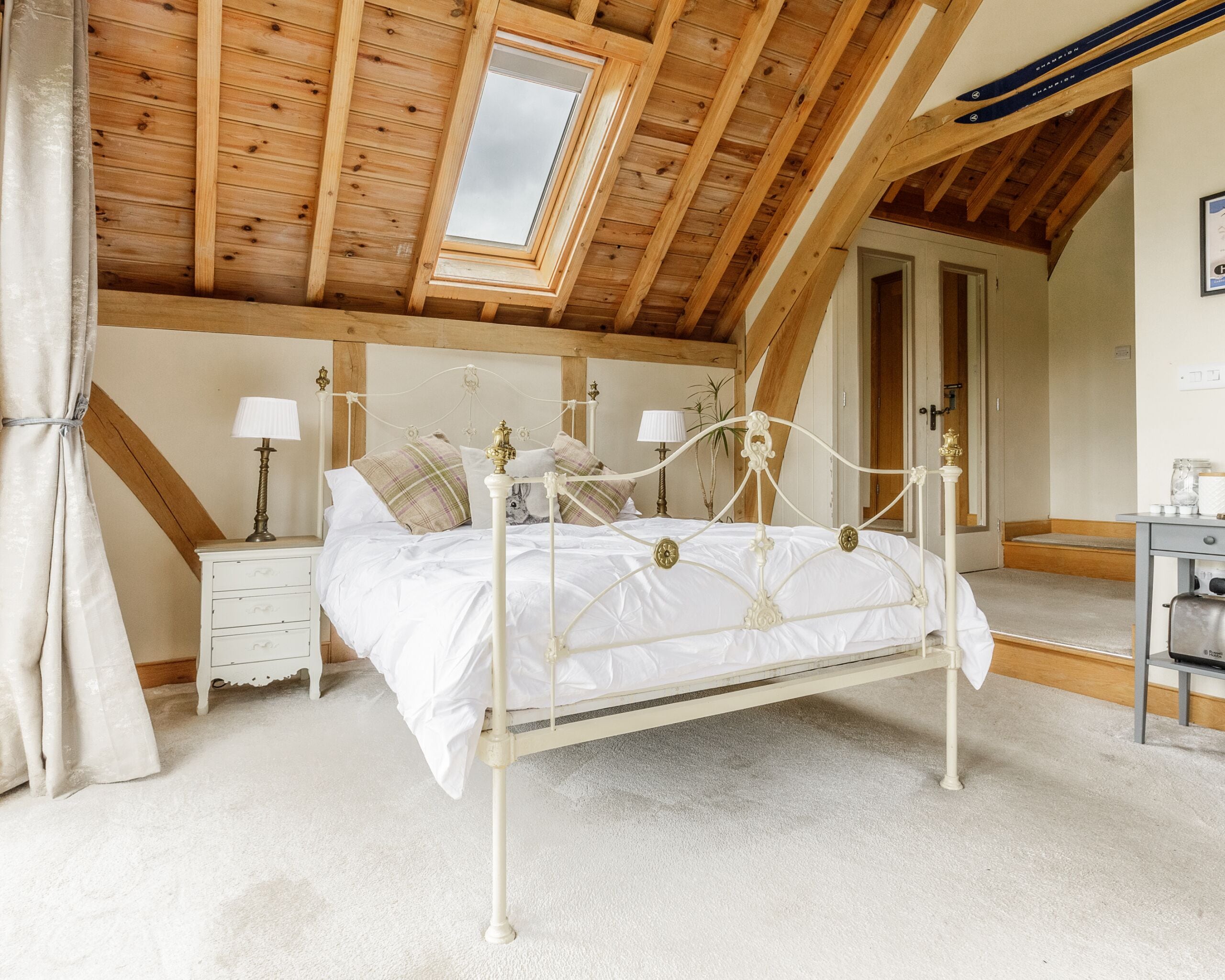 A bedroom with a white four-poster bed, a small bedside table with a lamp, a wooden ceiling with a skylight, and a door leading to another room.