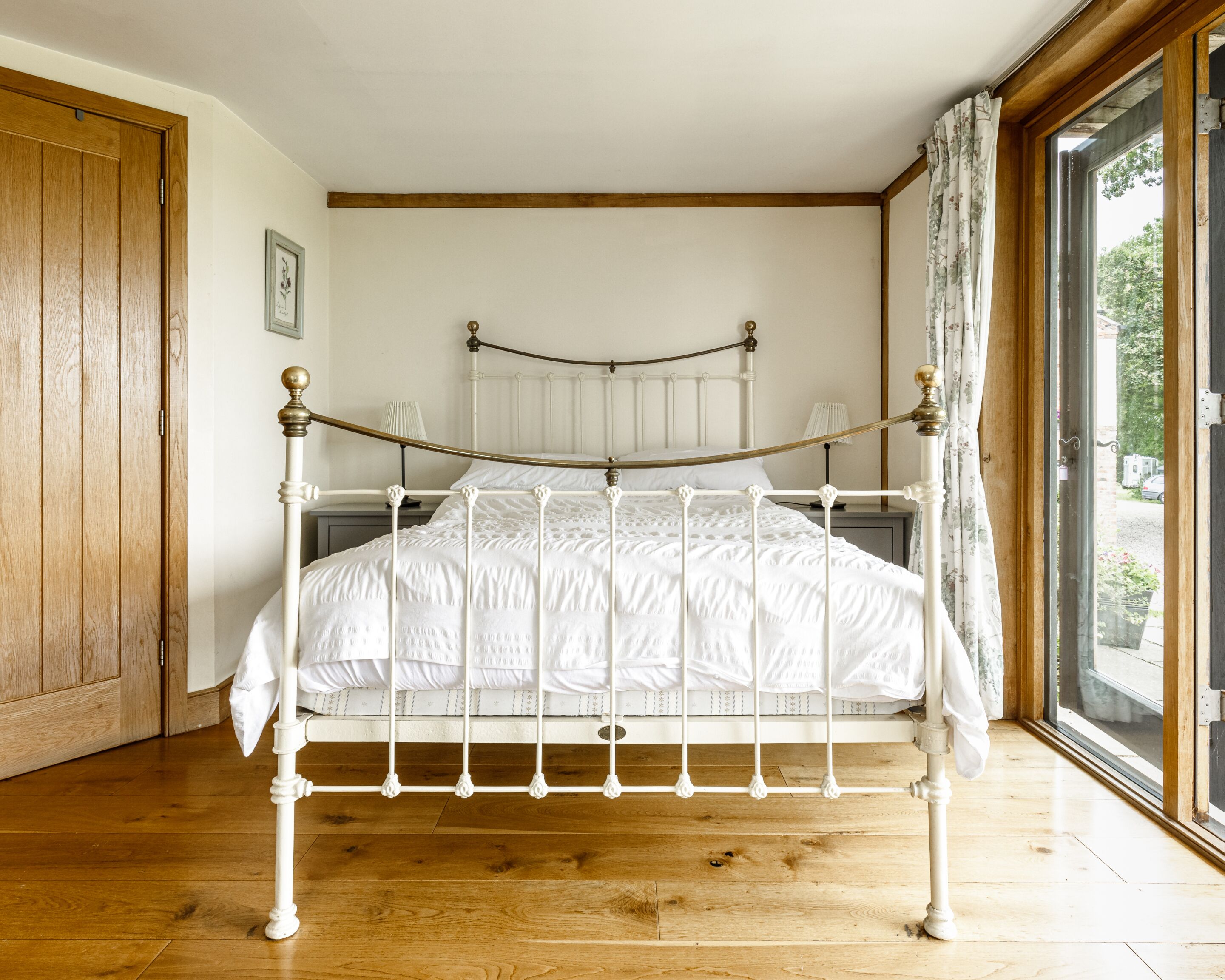 Una habitación con una cama de estructura metálica blanca, suelo de madera, una puerta de madera y una ventana con cortinas.