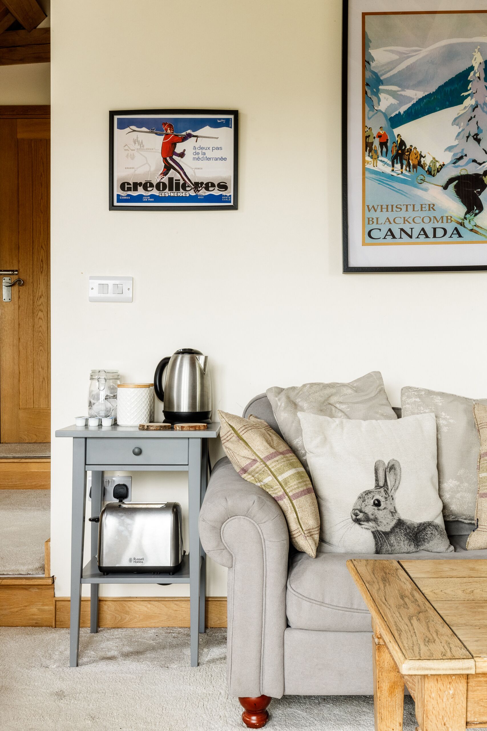 A cozy living room with a grey sofa, a wooden coffee table, and a small side table with a kettle and a toaster. There are pillows on the sofa, and a framed picture of a skier on the wall.