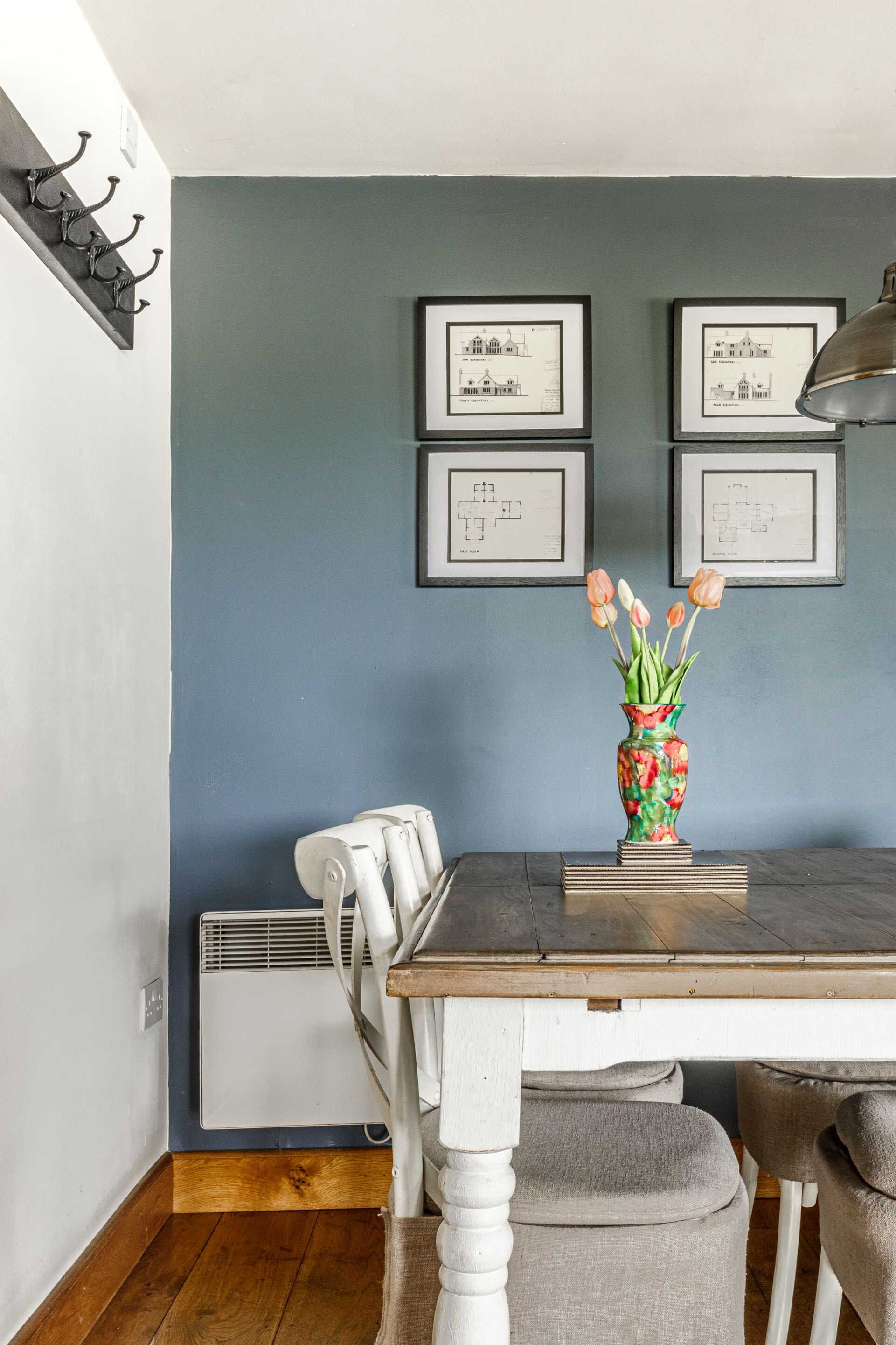 Un comedor con una mesa, sillas, un jarrón con flores y dibujos arquitectónicos enmarcados en la pared.