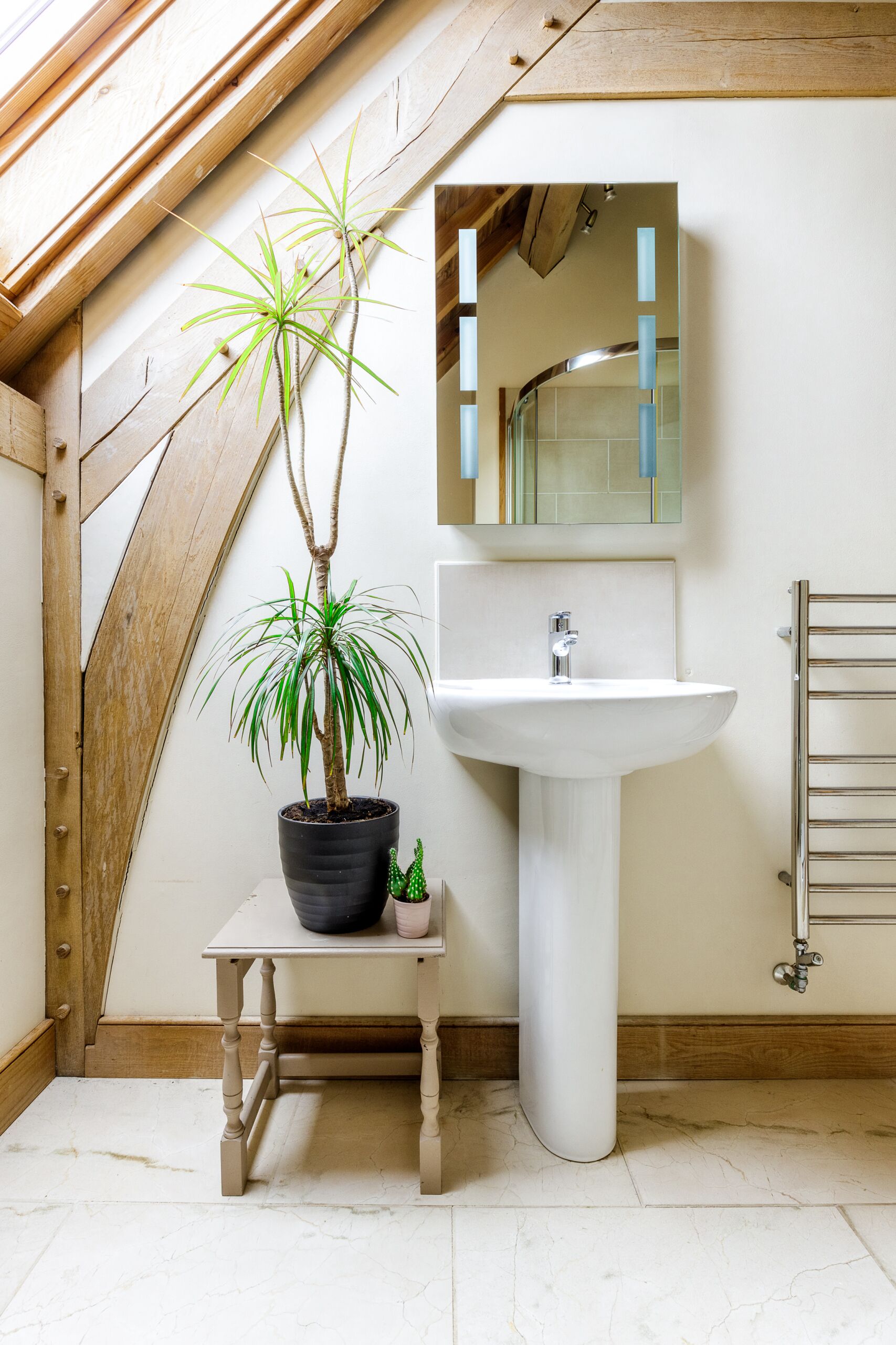A bathroom with a white pedestal sink, a wooden bench, a potted plant, and a wooden ceiling beam.