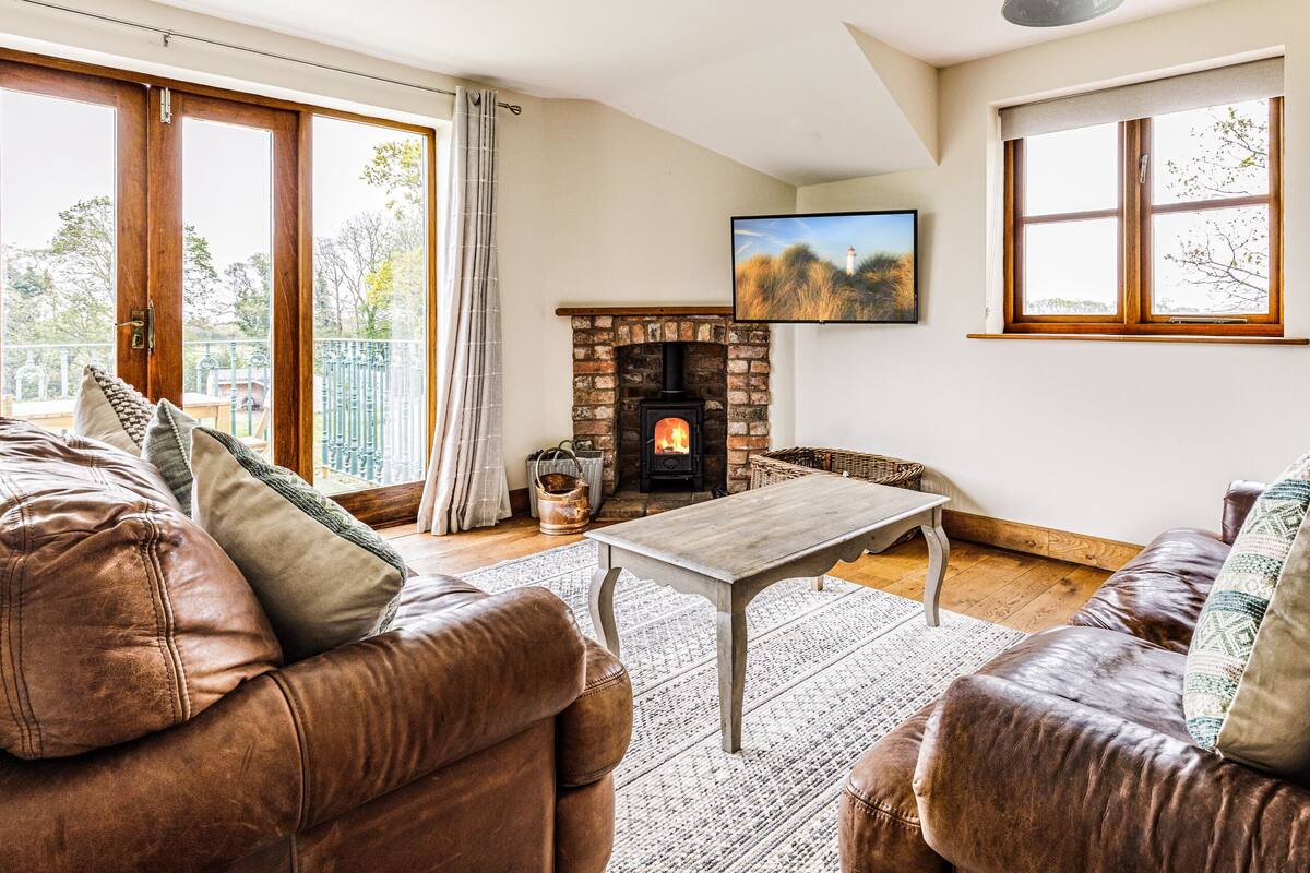 A cozy living room with leather sofas, a fireplace, and large windows offering a view of the outdoors.