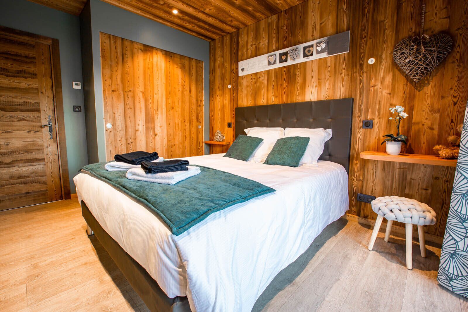 A neatly made bed with a teal blanket and pillows, a wooden headboard, a small stool, and a shelf with a potted plant.