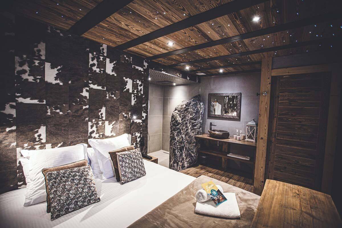 A modern bedroom with a wooden ceiling, a large bed with patterned pillows, a wooden nightstand, and a small table with a towel and book.
