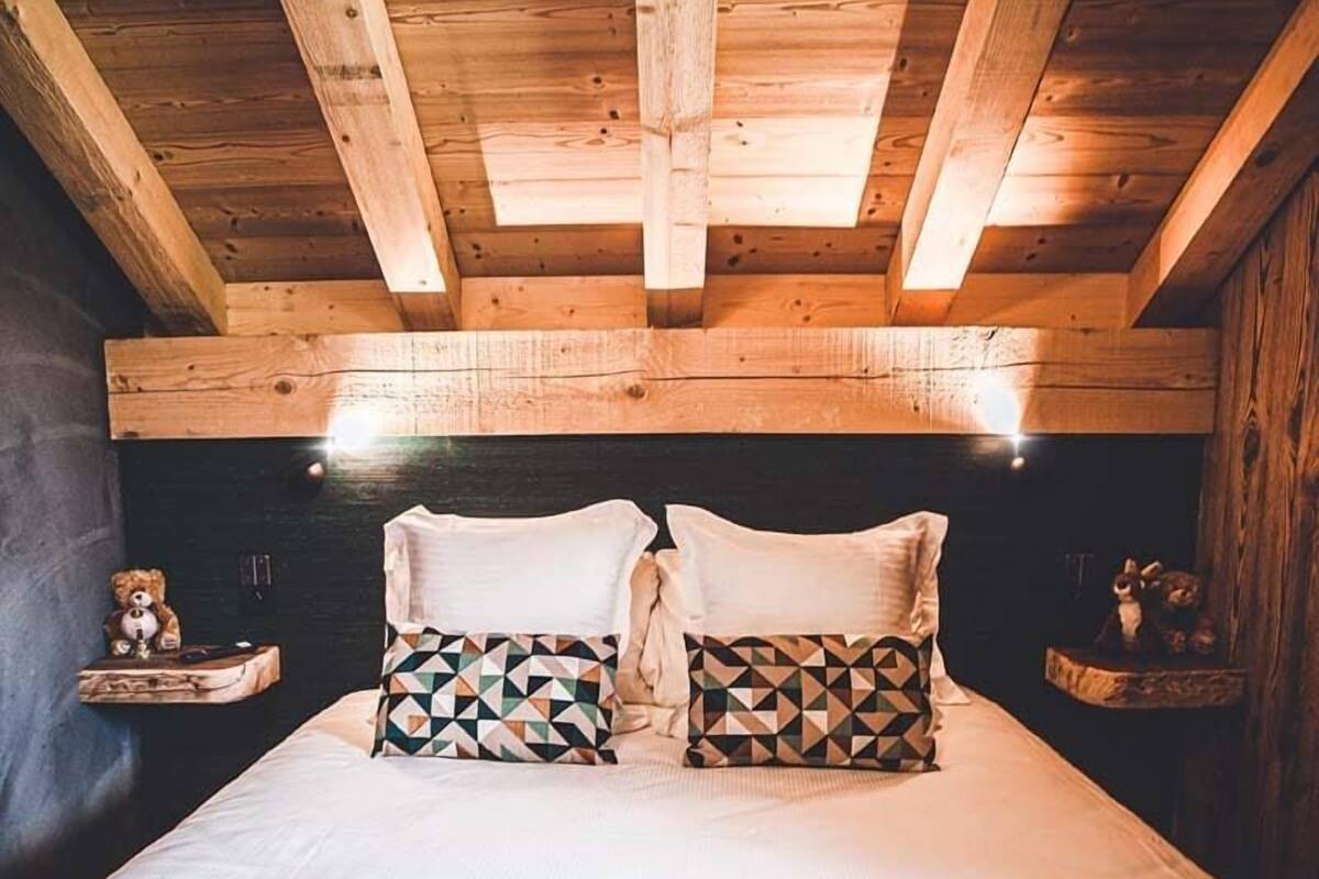 A wooden ceiling with exposed beams, a bed with patterned pillows, and a nightstand with a teddy bear.