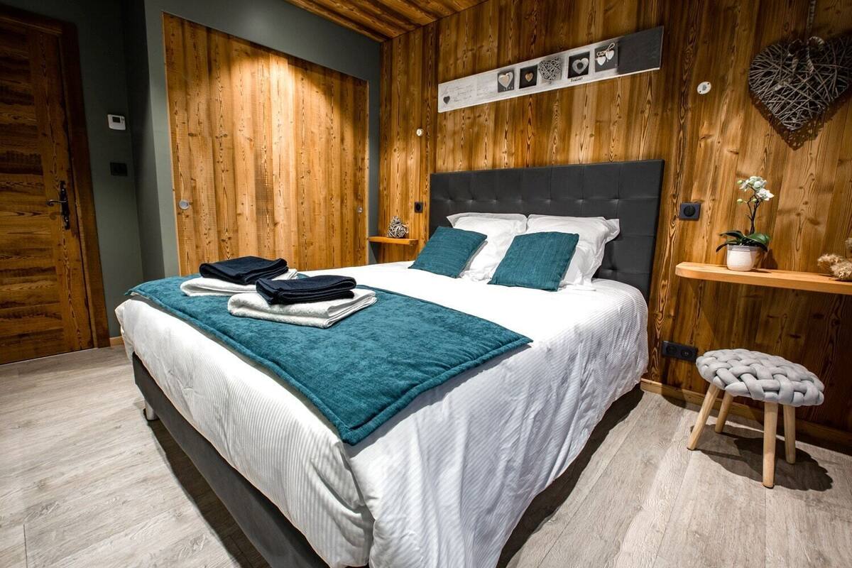 A modern bedroom with a wooden wall panel, a bed with white bedding and teal pillows, a small stool, and a shelf with decorative items.