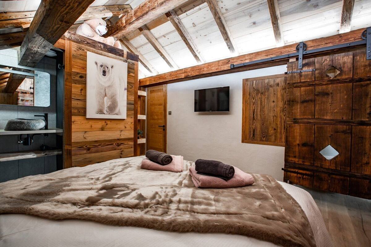 A cozy room with a wooden ceiling, a bed with a fluffy blanket, a mounted TV, and a wooden wardrobe.