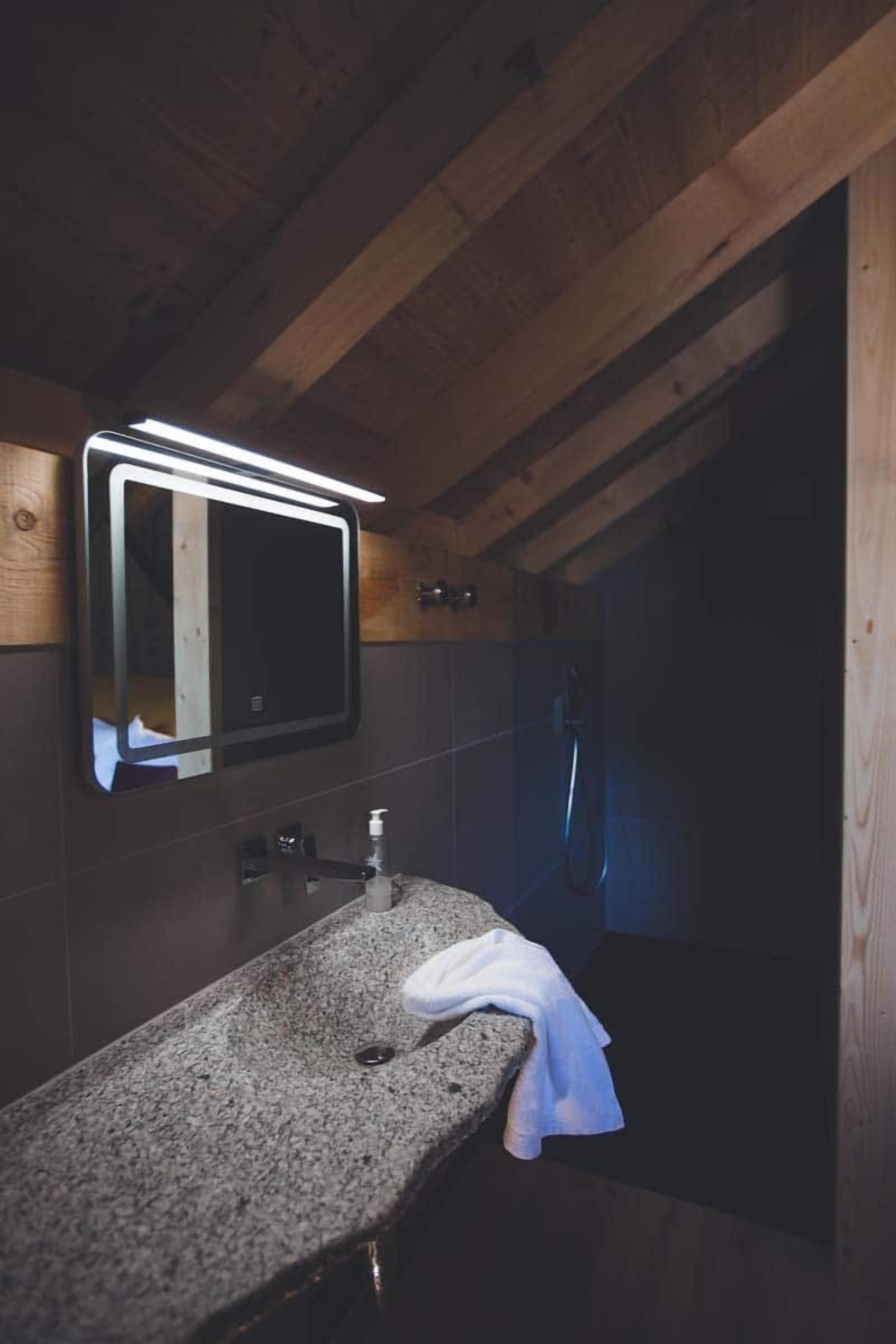 Un baño con encimera de granito, un espejo con iluminación integrada, un cabezal de ducha manual y una toalla sobre la encimera.