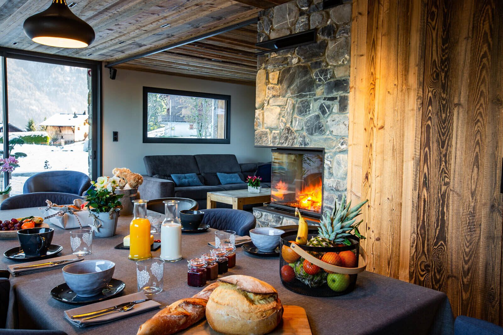 Ein gemütlicher Essbereich mit Kamin aus Stein, einem Tisch, der mit Frühstücksangeboten gedeckt ist, und Blick ins Freie.