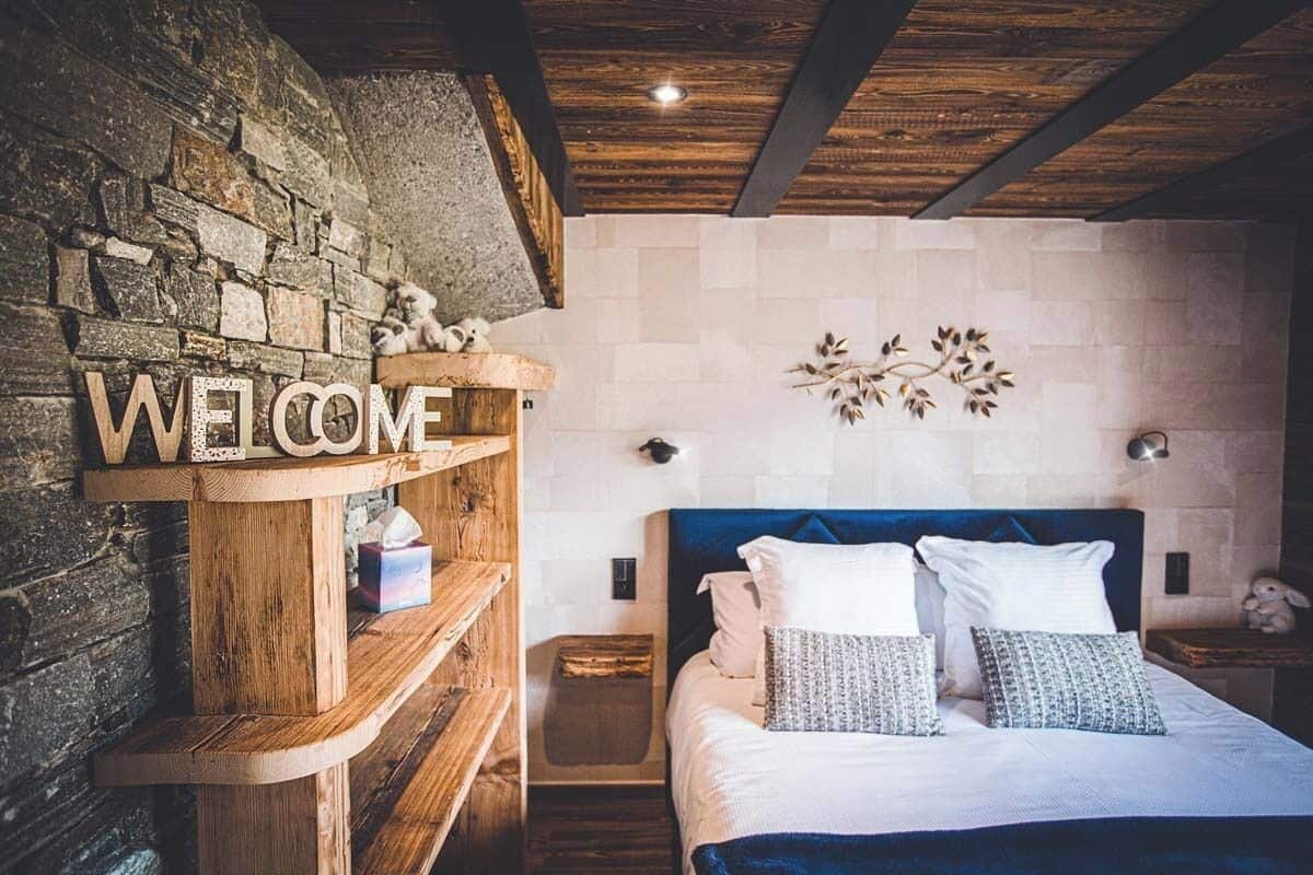 A cozy bedroom with a stone wall, a wooden shelf with a 'WELCOME' sign, a bed with white and patterned pillows, and a blue headboard.