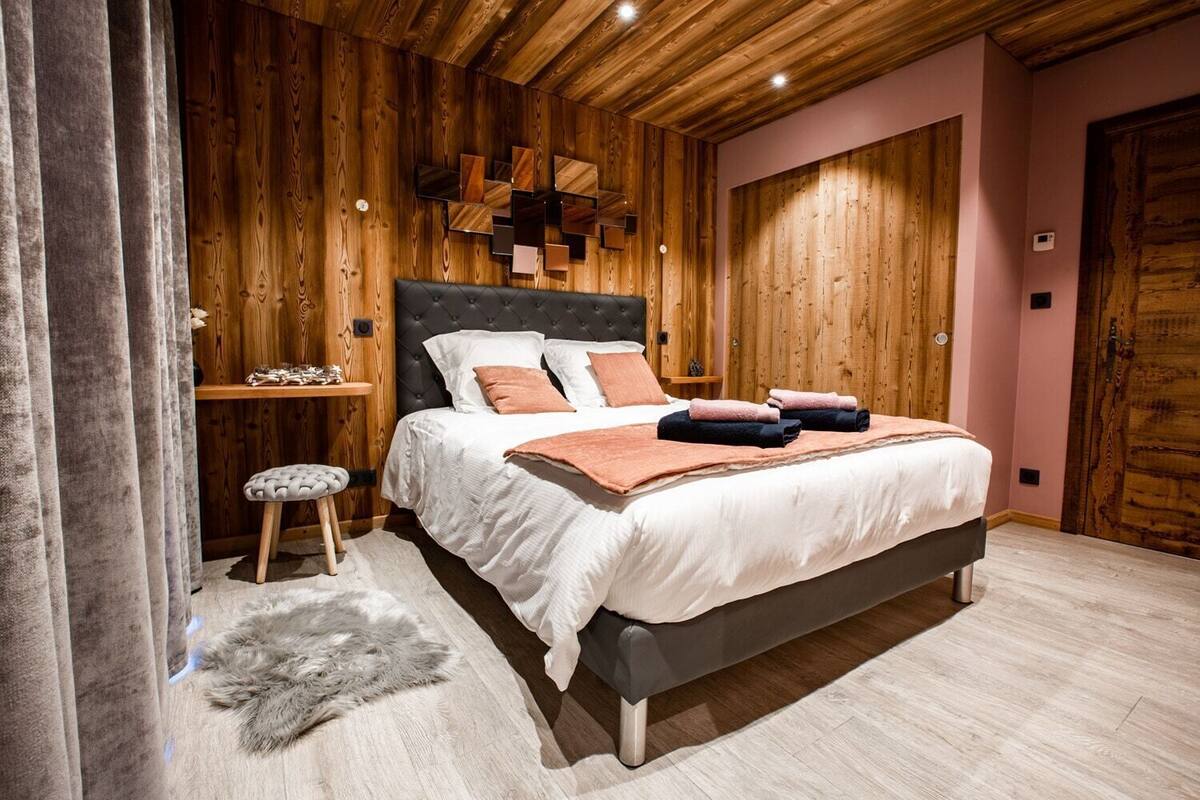 A bedroom with a wooden ceiling, a tufted headboard, a bed with white and pink bedding, a small stool, and a fur rug.