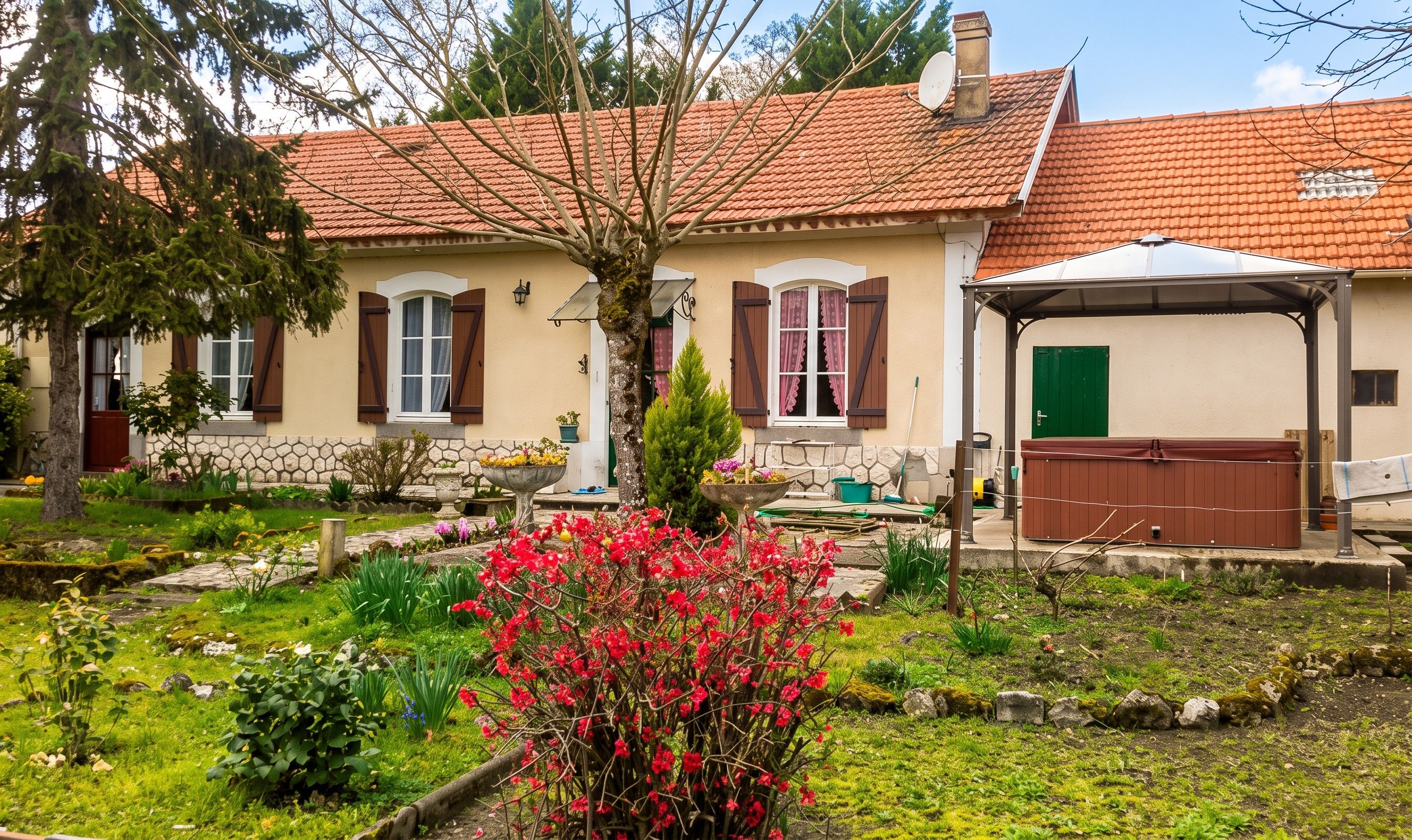 Een huis met een rode dakbedekking, een tuin met verschillende planten en bloemen, en een groene deur.