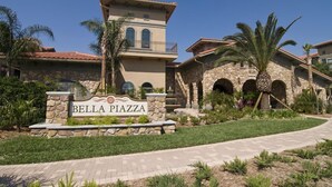 Une enseigne pour Bella Piazza, avec une façade en pierre et des fenêtres en arc de cercle, le tout entouré de palmiers et d’une pelouse bien entretenue.