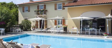 A swimming pool with lounge chairs and umbrellas, surrounded by a building with a tiled roof and windows with shutters.