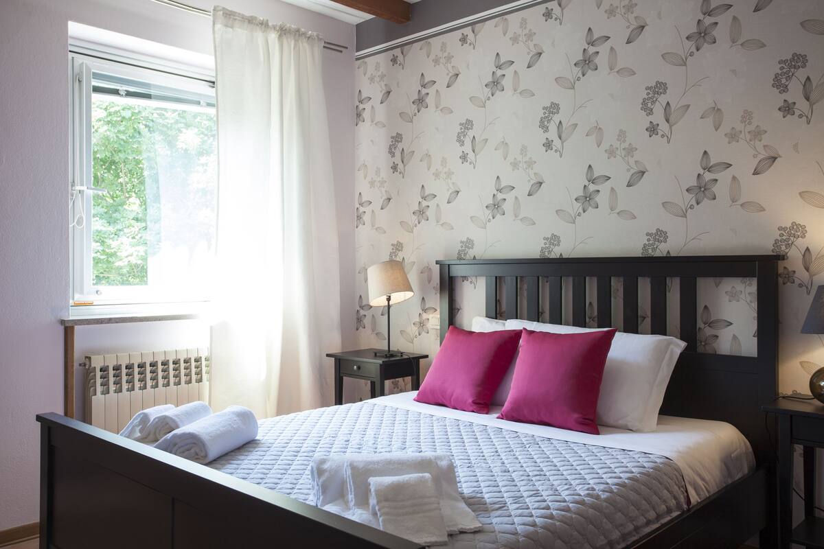 Una cama bien tendida con almohadas blancas y rosas, una mesita de noche con lámpara y un estampado floral en el papel tapiz.