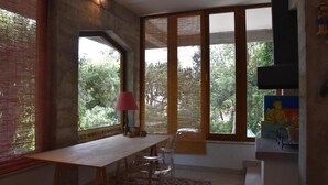 Une pièce avec une table et des chaises en bois, de grandes fenêtres équipées de stores en bambou et un téléviseur fixé au mur.
