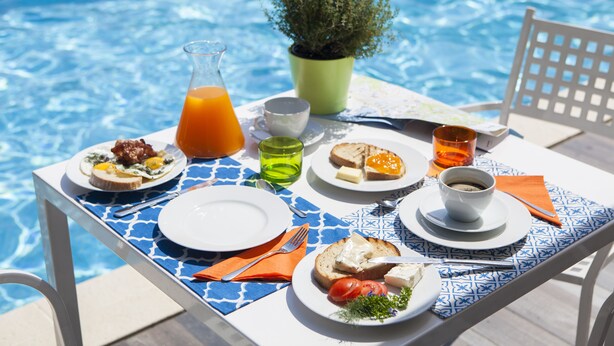 Une table à manger placée près d’une piscine, avec des aliments pour le petit-déjeuner, un pichet de jus d’orange et une plante en pot.