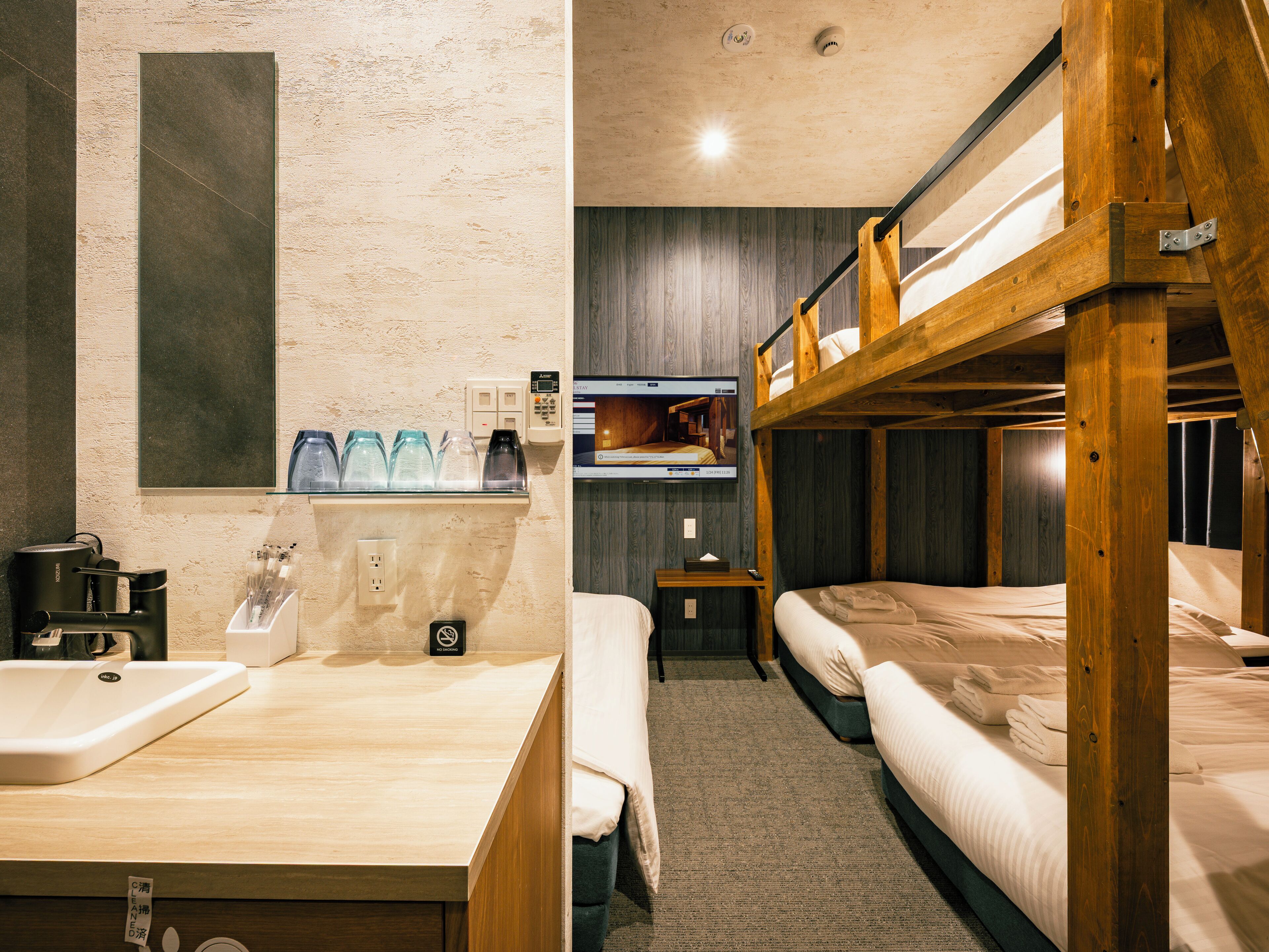 A hotel room with a wooden bunk bed, a bathroom with a sink and mirror, and a television on the wall.