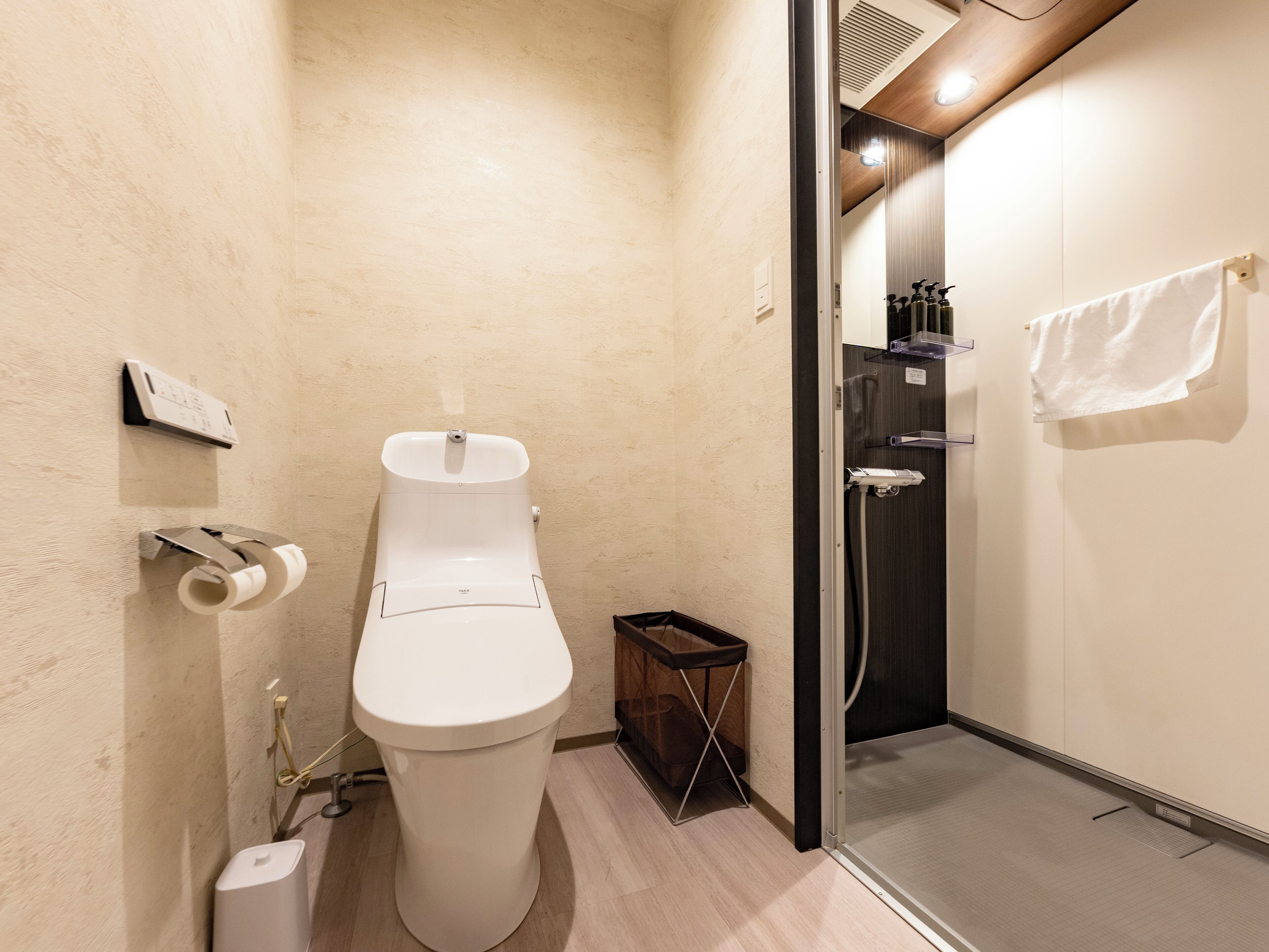 A modern bathroom with a toilet, a paper towel dispenser, a small trash bin, and a shower area.
