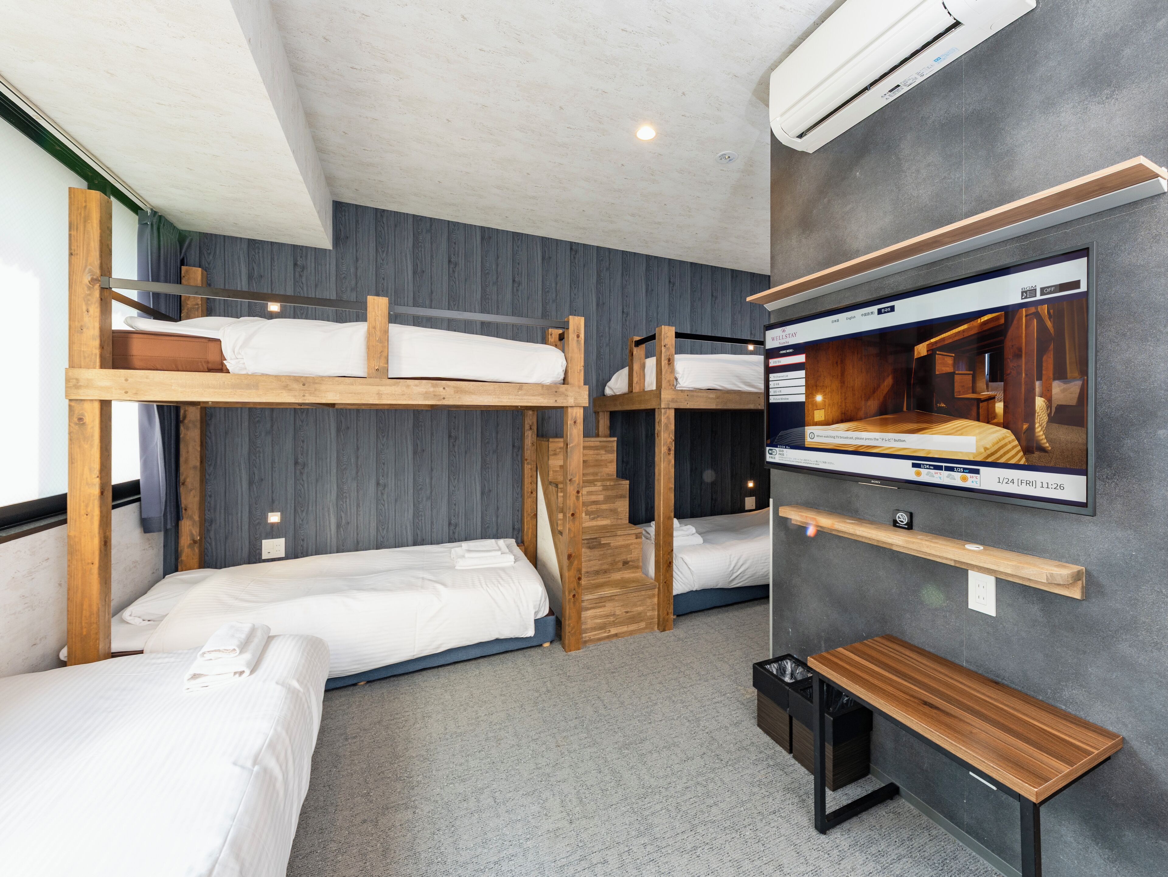 A room with bunk beds, a TV mounted on the wall, a wooden bench, and a trash bin.