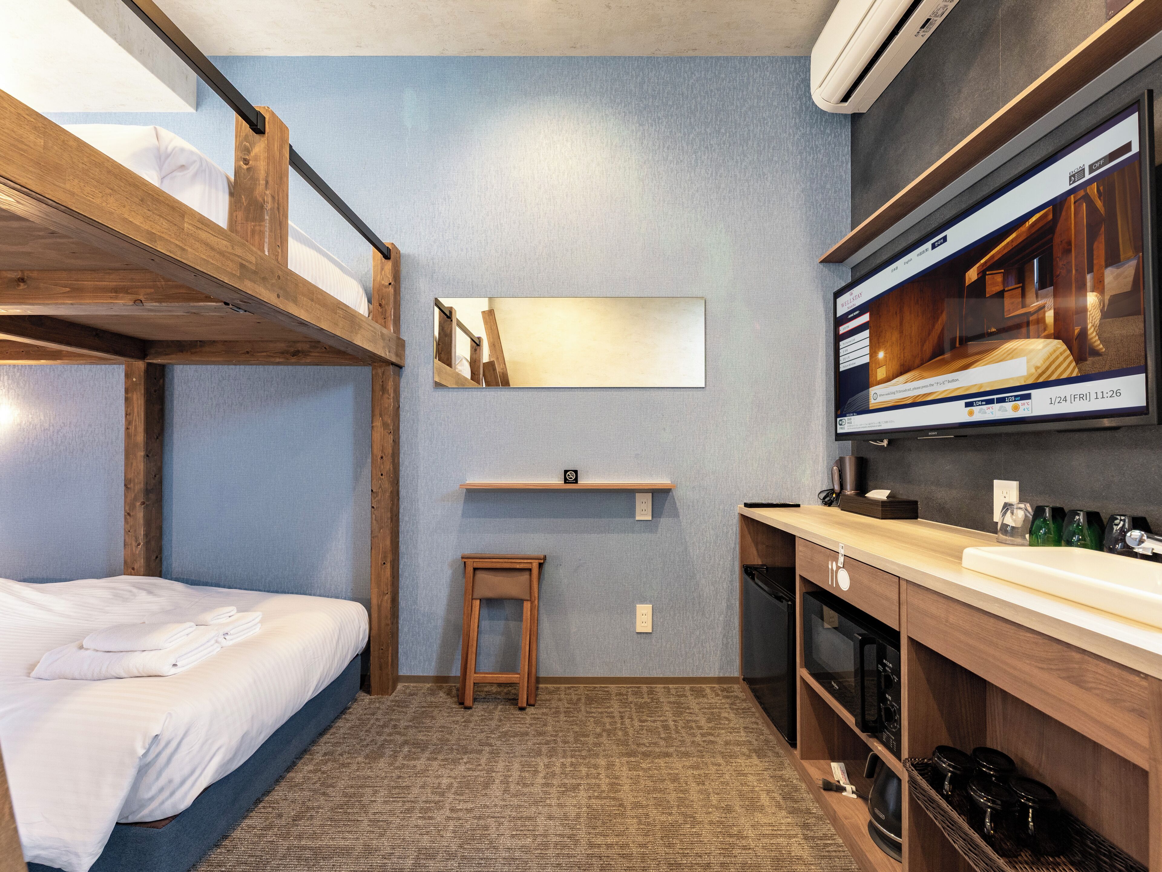 A compact hotel room with bunk beds, a small stool, a mirror, a TV, a microwave, and a sink.