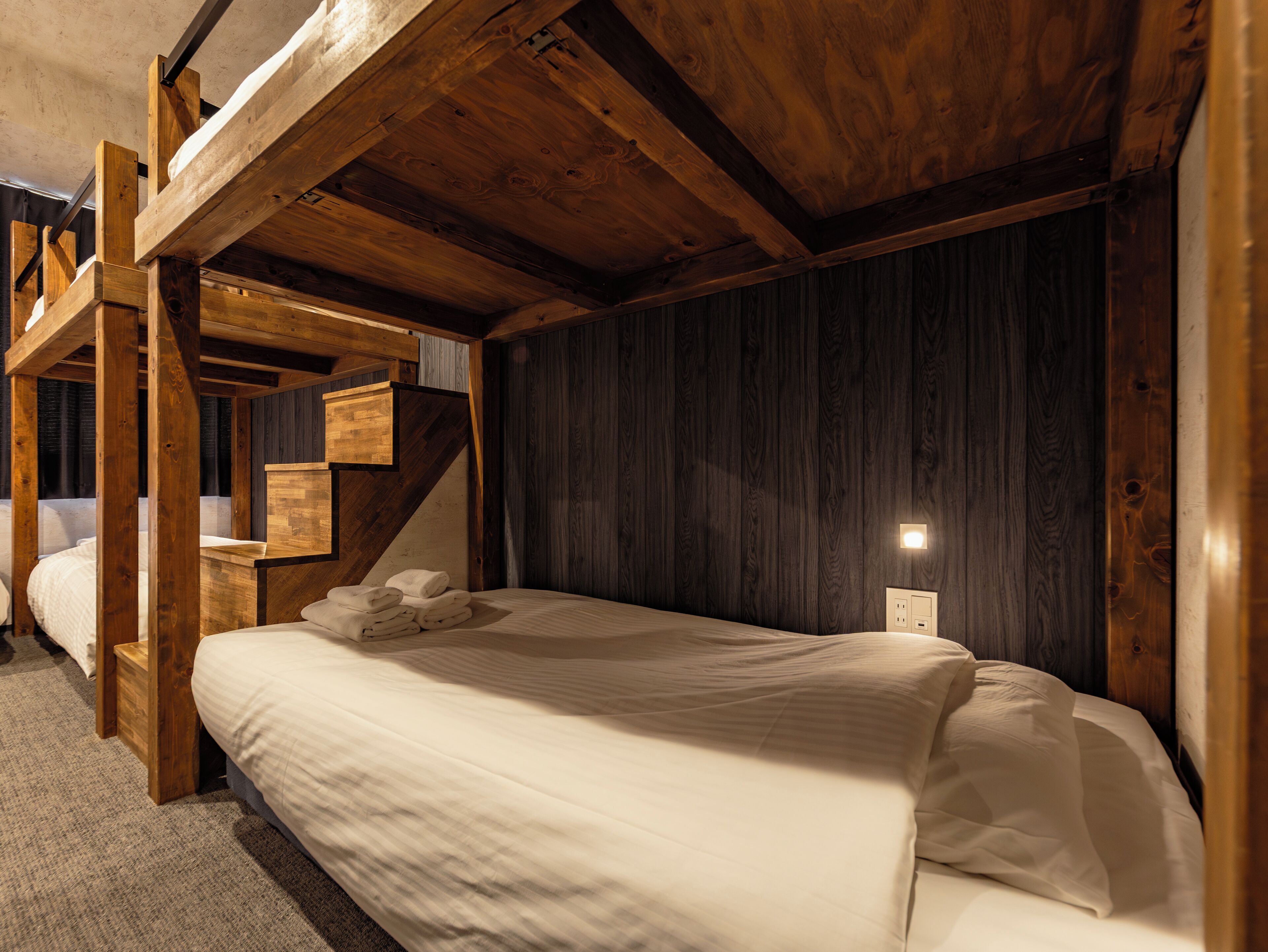 A wooden room with a bunk bed, a bedside table, and a wall-mounted light.