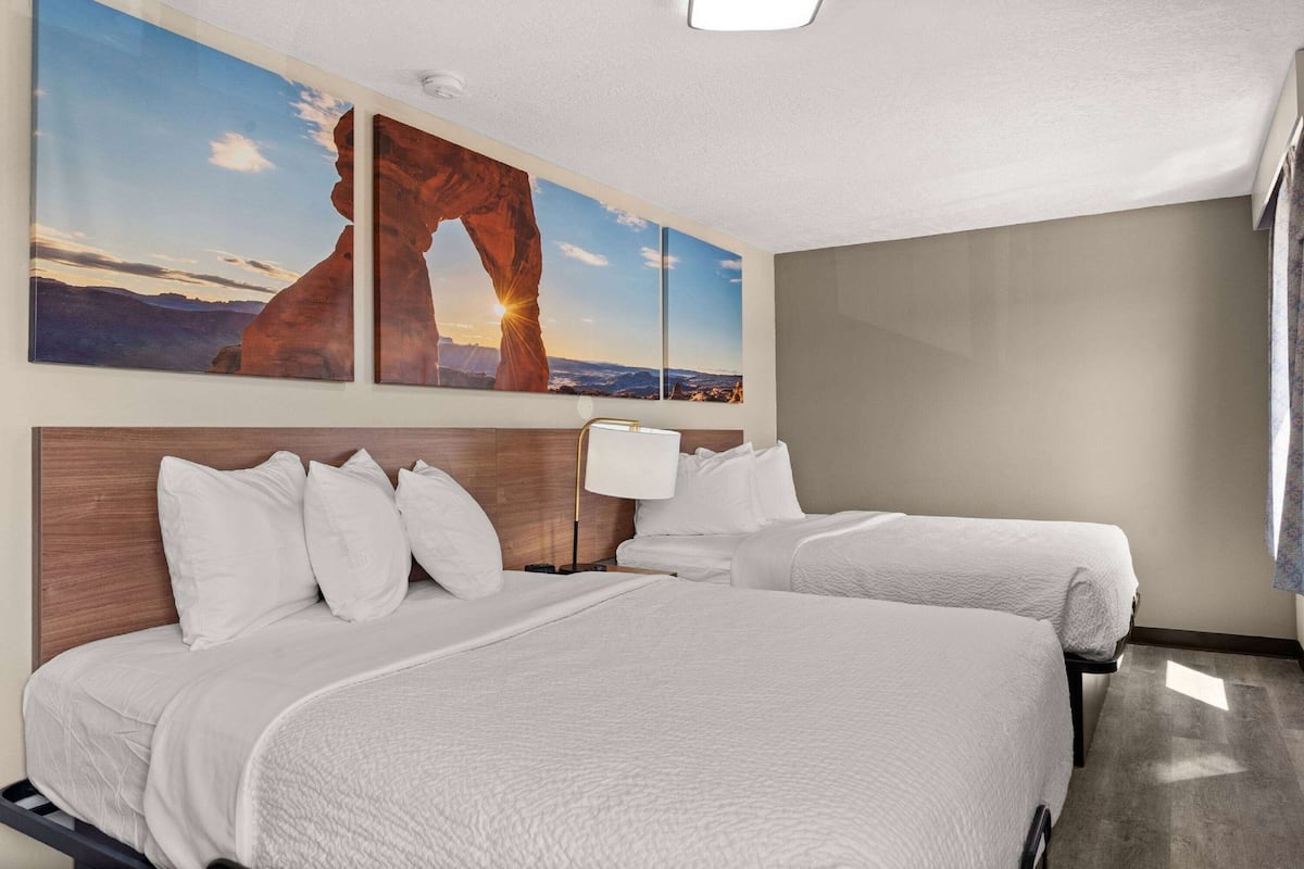 A hotel room with two beds, a wooden headboard, a nightstand, a lamp, and a large wall painting of a rock formation.