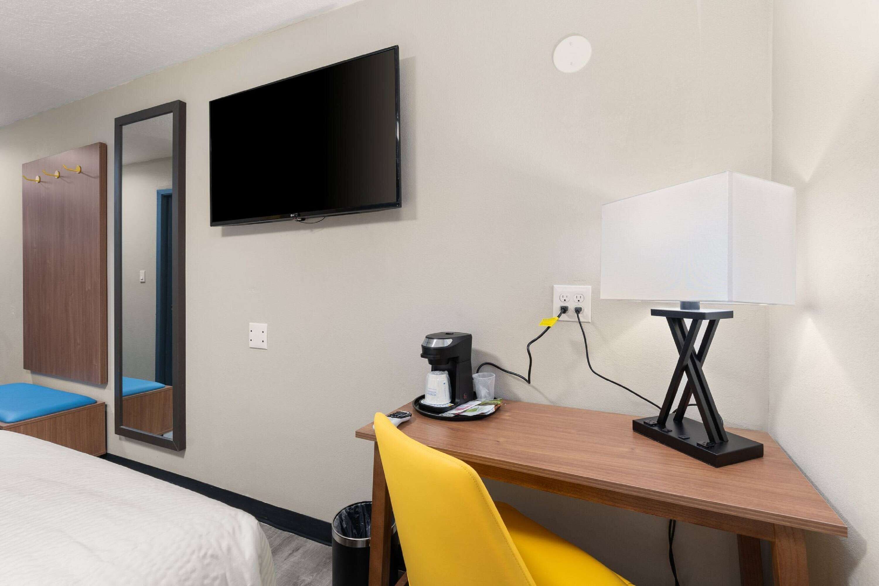 A hotel room with a bed, a desk with a lamp, a coffee maker, and a TV on the wall.
