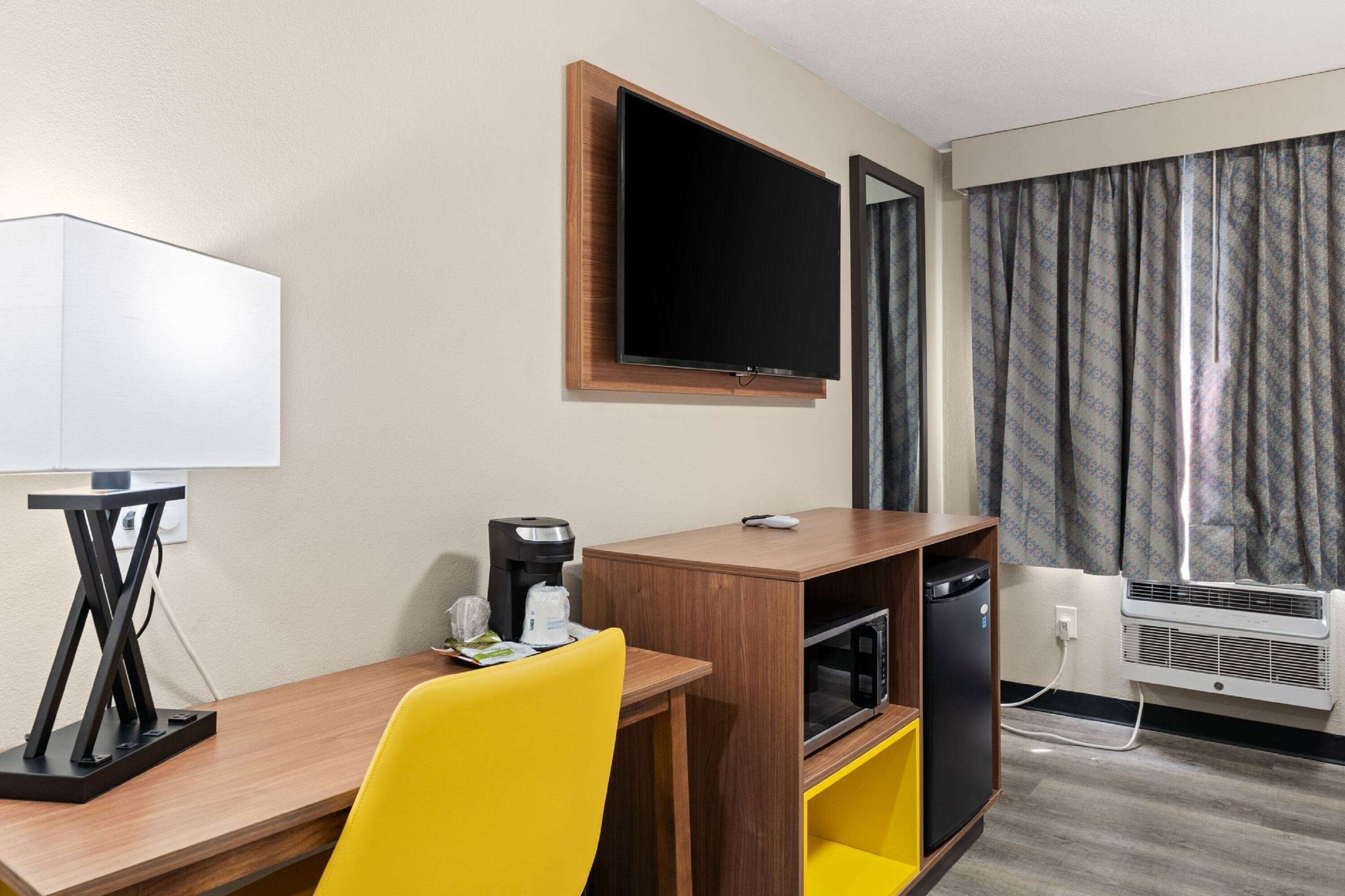 A hotel room with a wooden desk, a television mounted on the wall, a coffee maker, a microwave, and an air conditioning unit.
