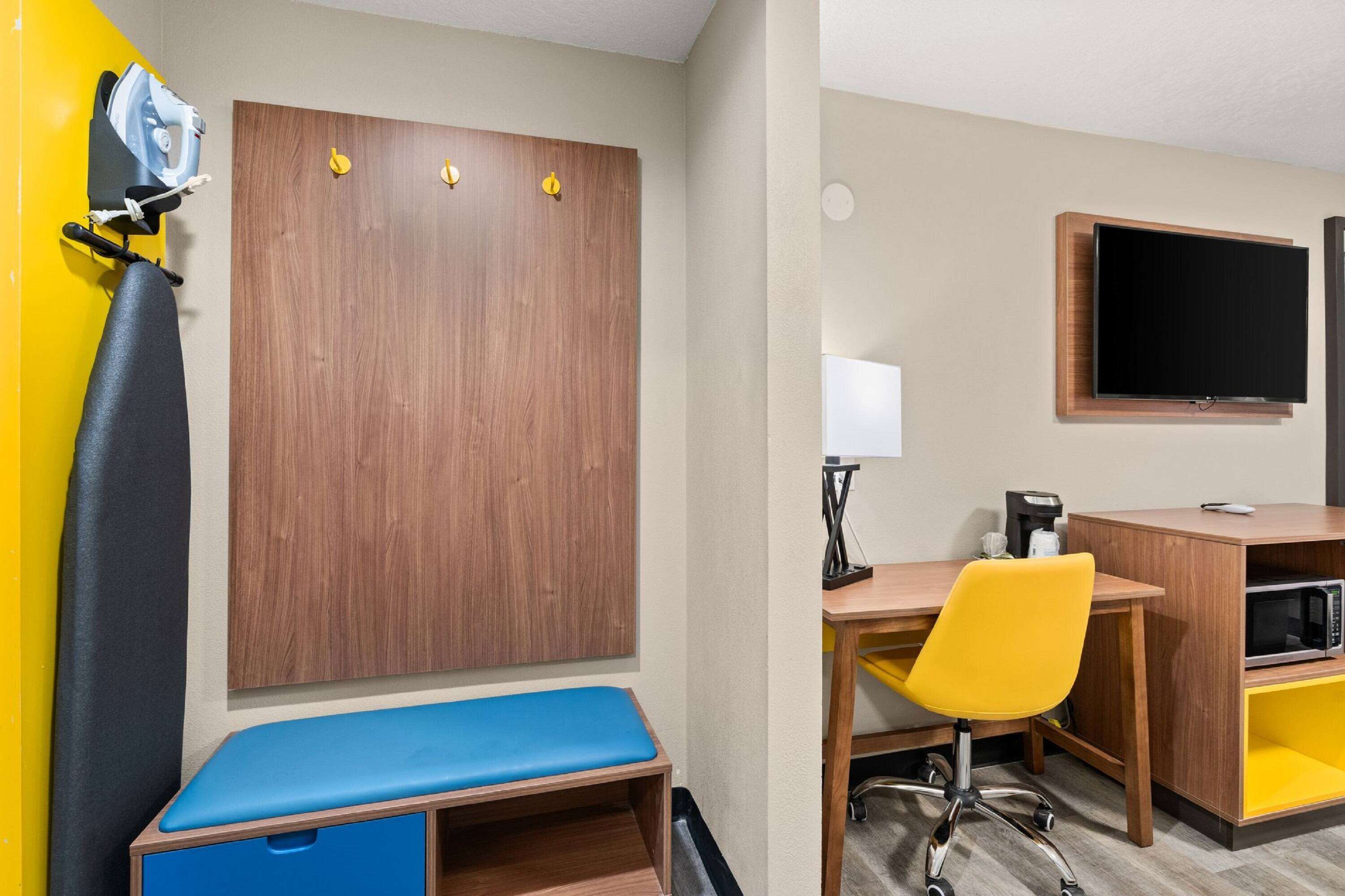 A hotel room with a wooden wardrobe, a blue bench, a desk with a yellow chair, a flat-screen TV, and a microwave.