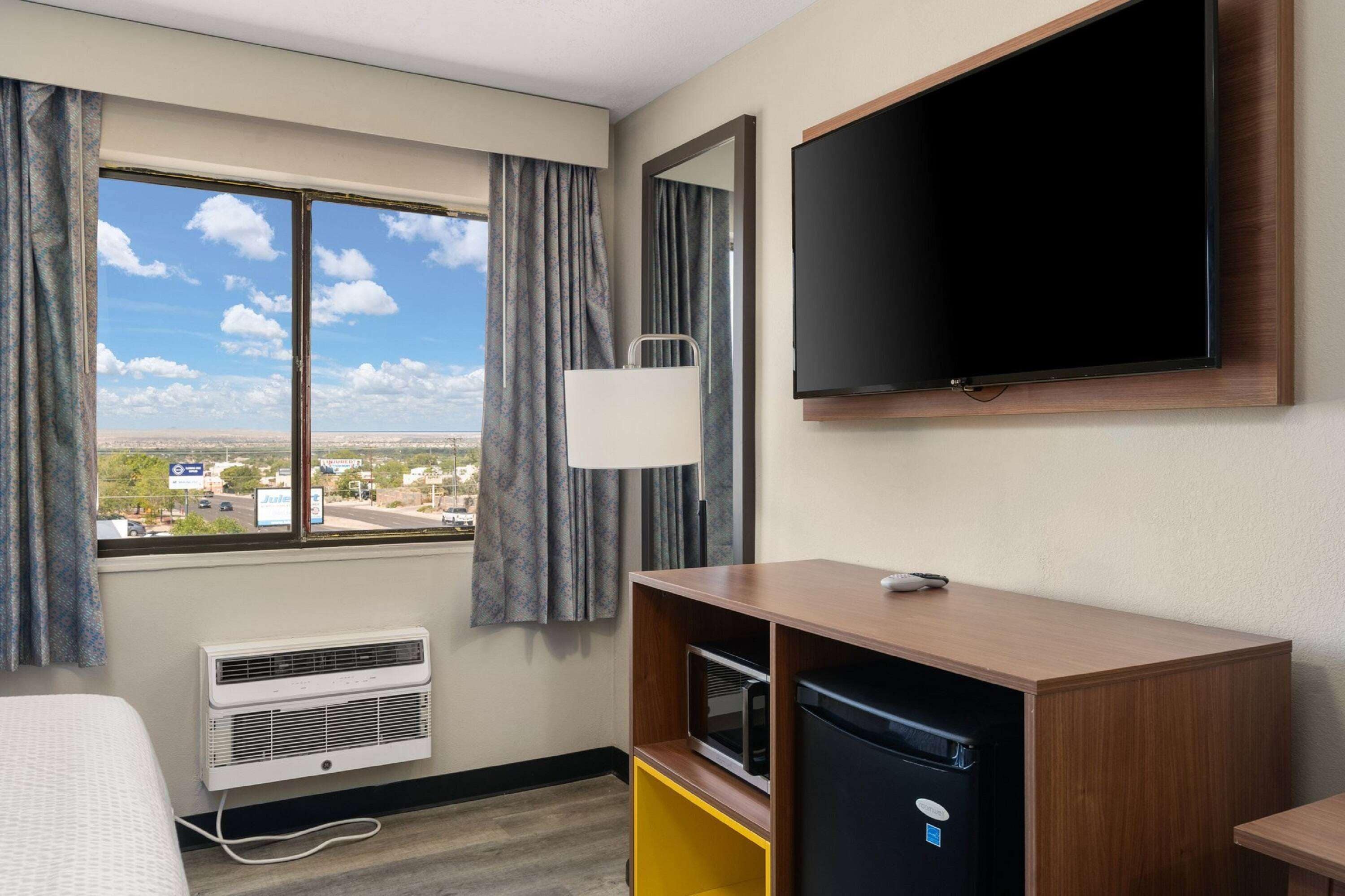 A hotel room with a large window, a flat-screen TV mounted on the wall, a small refrigerator, and a bedside table.