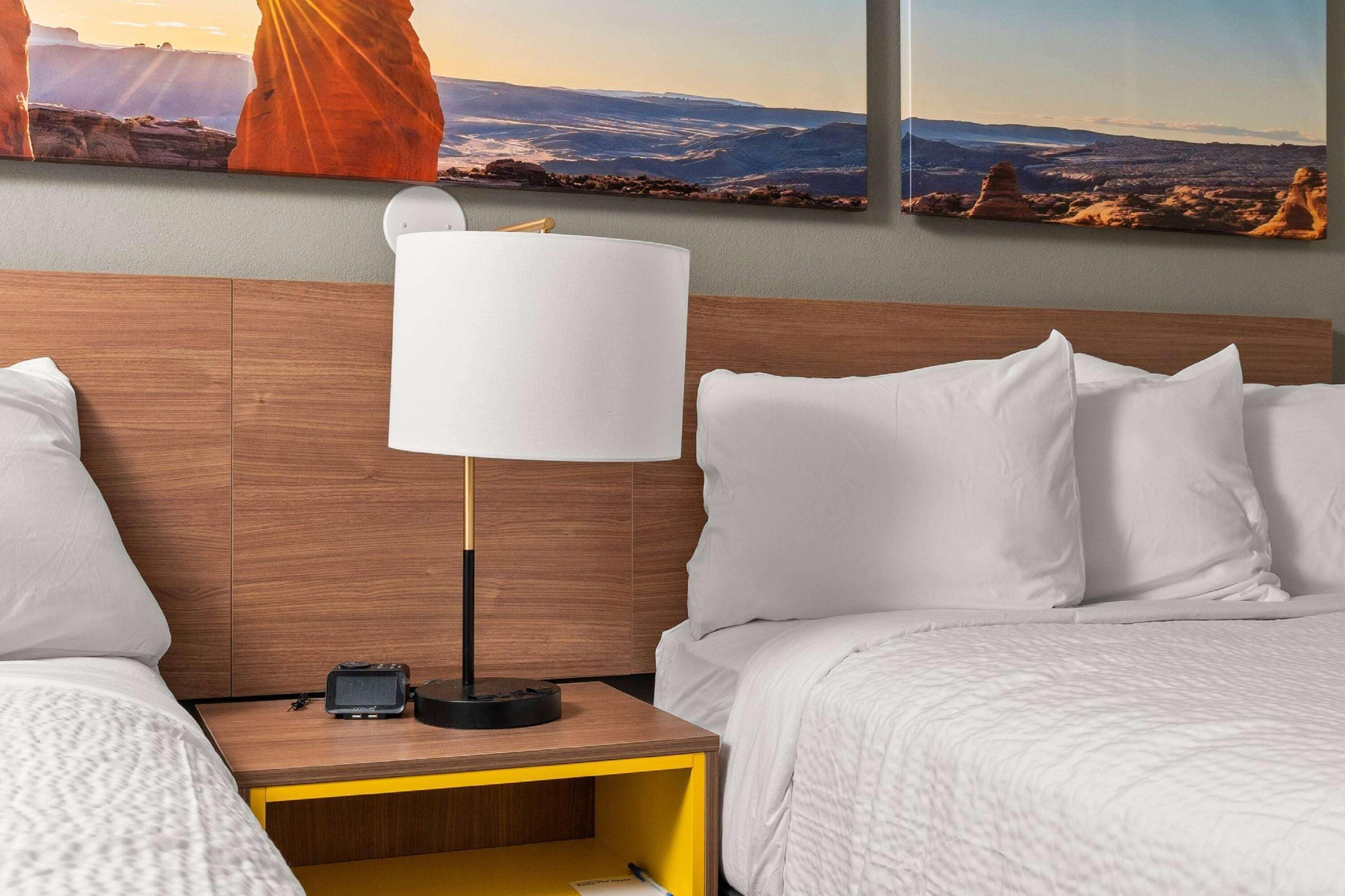 Habitación de hotel con cabecera de madera, ropa de cama blanca, mesita de noche con lámpara y un cuadro de paisaje sobre la cama.