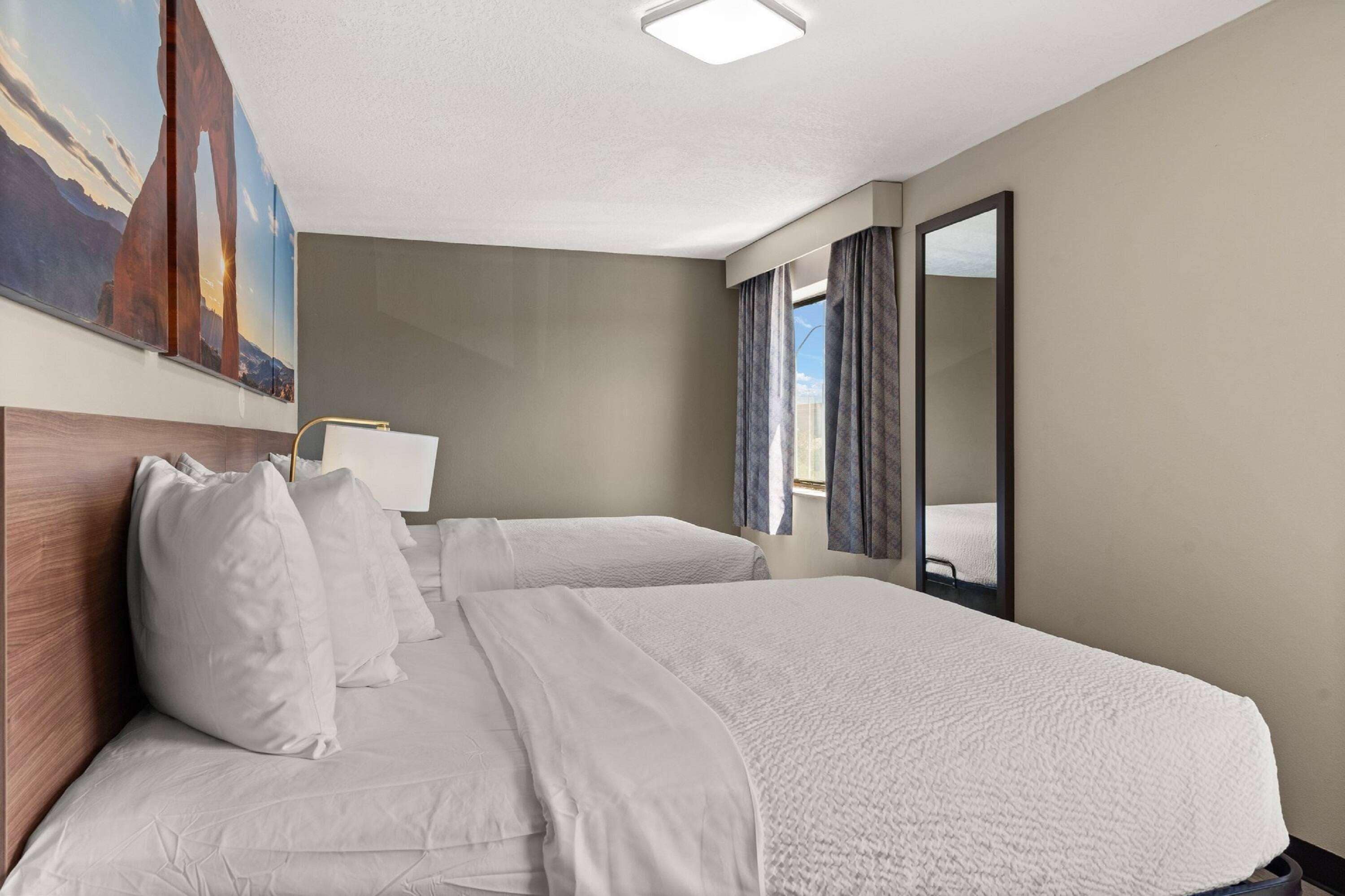 Une chambre d’hôtel avec deux lits, une grande fenêtre avec des rideaux, un tableau représentant un pont et un luminaire au plafond.